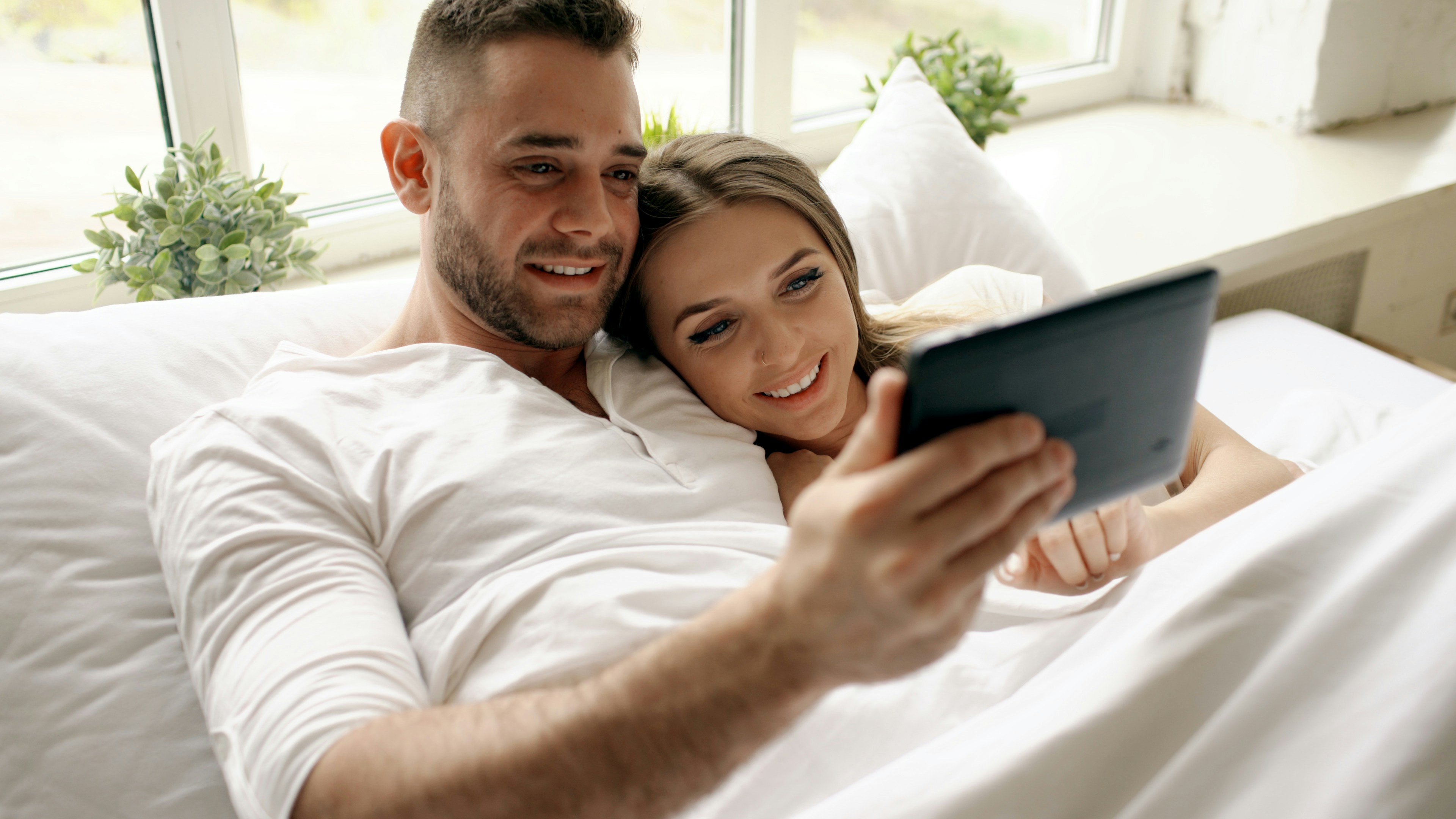 Young cute and loving couple using tablet and talking lying in bed in bedroom at morning